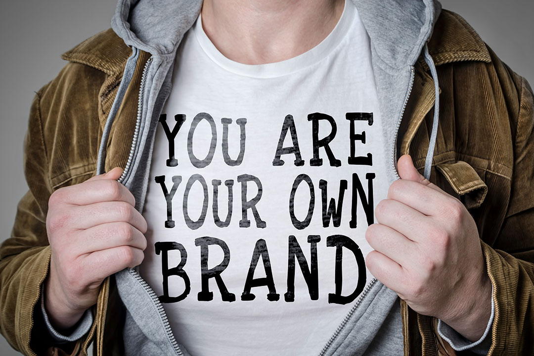 A person spreading open their jacket layers to reveal a white shirt that says “You are your own brand.”