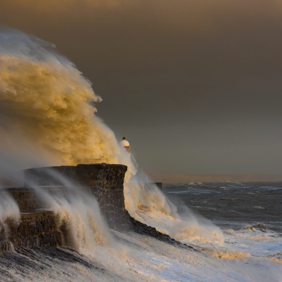 A huge ocean wave crashing onto the stone path that leads to a lighthouse