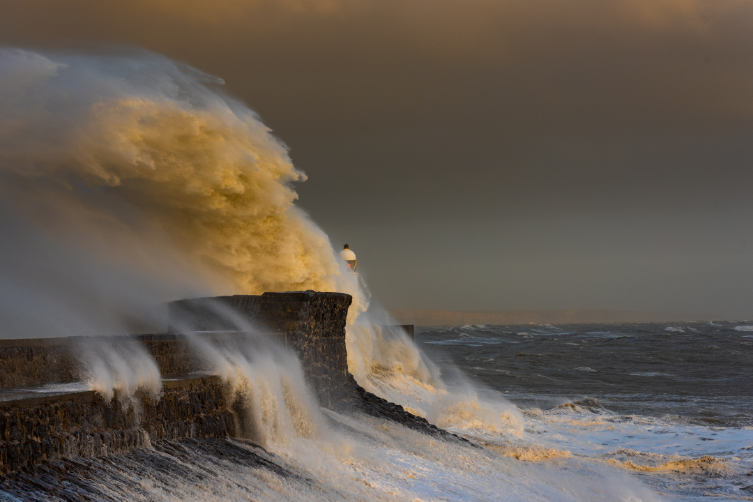 A huge ocean wave crashing onto the stone path that leads to a lighthouse