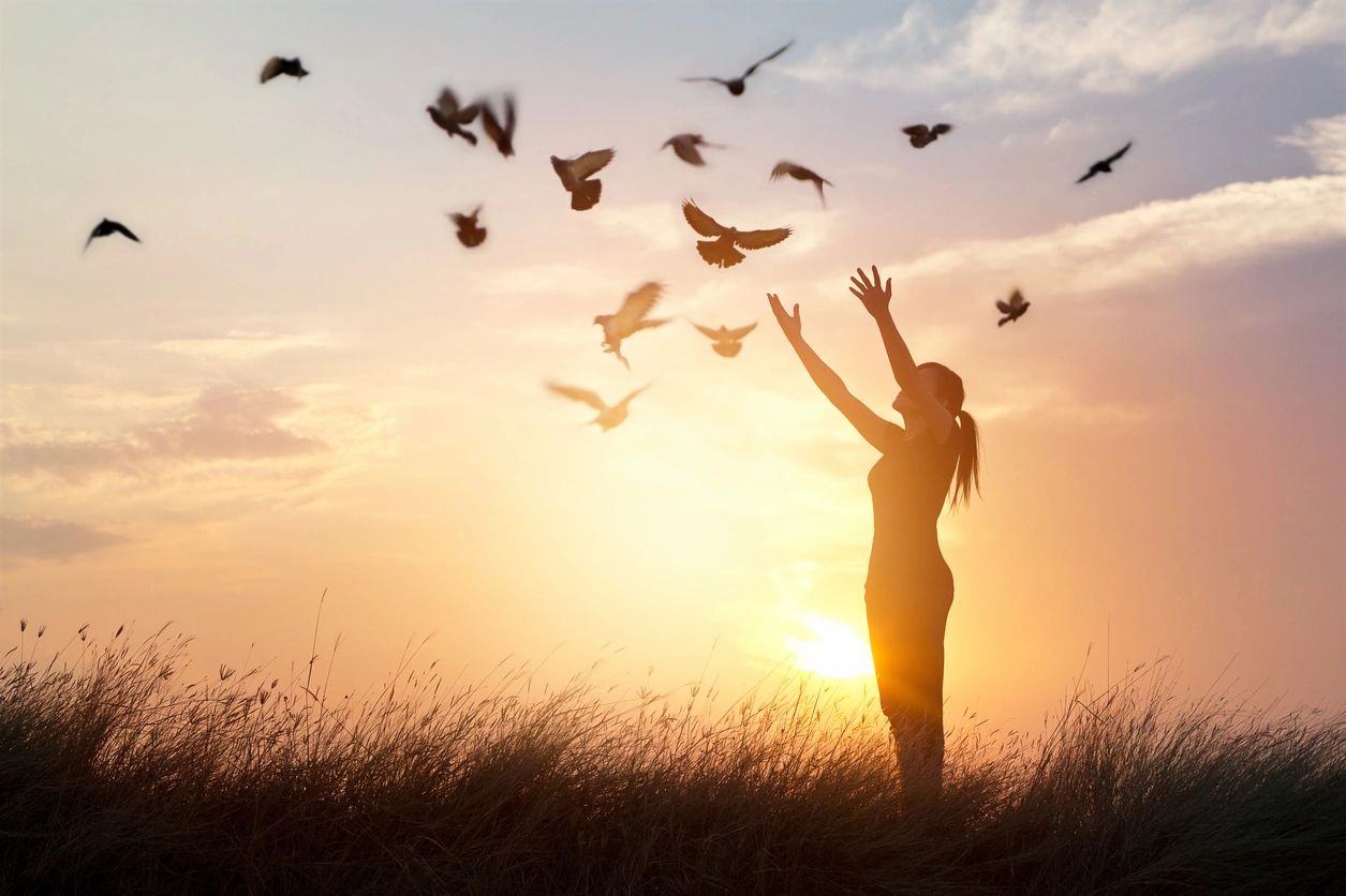 Woman in a field raising her arms to feel a flock of birds during sunset